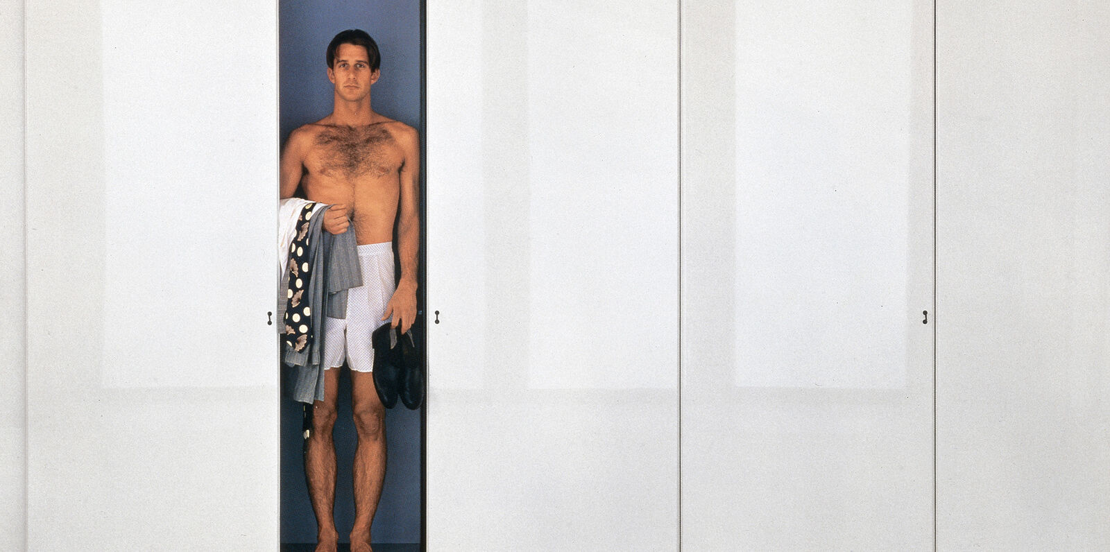 Sisamo Wardrobe. A man in white pants. On his right arm clothes, holding his shoes.